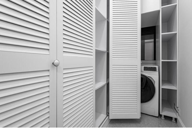 White louvred wardrobe doors and open shelving beside a front-loading washing machine